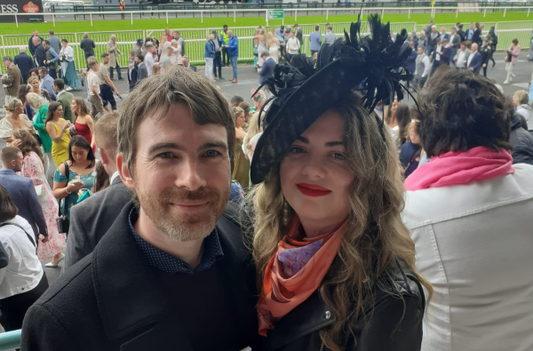 Irish scarf designer at Galway races with boyfriend wearing a peach luxury silk watching horse racing and with feather hat. Accessories and fashion print.  